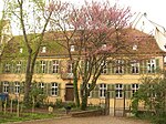 Photographie d'un parc public avec des arbres devant un bâtiment jaune de deux étages avec des volets verts.