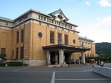 In front of a brick and concrete building, with driveway to a covered entrance, and a curved Japanese gable