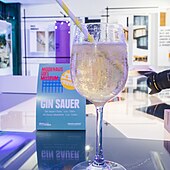 Ein sprudelndes Kaltgetränk im Stielglas mit Strohhalm, dahinter eine improvisierte Ausstellung und ein Aufsteller „Modehaus goes Museum“ und „Gin Sauer“.