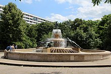 Großes rundes steinernes Wasserbecken, in der Mitte ein Wasserspiel, auf dessen Rand übergroße Bronze-Statuen von Frauen in liegender/sich stützender Haltung, ganz oben eine stehende Frauenstatue aus Bronze