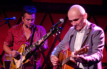 A man at left is playing an electric guitar, while 56-year-old Kelly plays his acoustic guitar. Both are looking down towards their own guitar, each uses a plectrum in their right hand while the left is on the fret board. In front of each is a microphone on its stand.