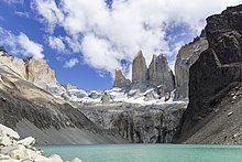 Un resserrement dans la vallée bloque un glacier et préserve un grand lac de montagne. Au-dessus trois pics neigeux volent dans les nuages.