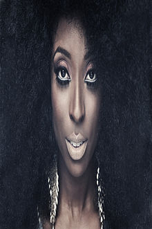 Face of a dark-skinned woman with big and curly black hair, looking at the camera, surrounded by darkness