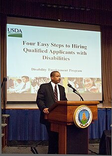 A man is speaking behind a microphone podium during a conference. Behind him, there is a screen showing a presentation slide reading "Four Simple Steps to Hiring Qualified Candidates with Disabilities"