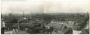 Photographie en noir et blanc de bâtiments et de rue vus en léger surplomb avec, à l'arrière-plan, un grand nombre de cheminées fumantes