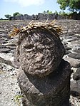 Weathered grey stone bust with a palm frond crown. Grey rocks with white and in between them in the background.