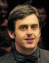 Headshot of a dark-haired man smiling at the camera, with black shirt, black waistcoat, and black bow tie visible