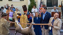 An older white man in a dark blue blazer smiles as he is presented with a dead deer hanging upside down held by two men in contemporary Native American attire.