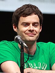 A man with brown hair wearing a green shirt smirks as he sits at a table in front of a microphone