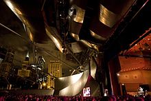 It is night. A spotlit lone performer plays an instrument on a large stage at right, and a large audience is visible across the bottom of the rest of the image. Large curving metal panels frame the stage and loom over the audience, while the lights of very tall skyscrapers shine against a cloudy sky behind.