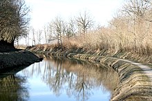 Photo en couleurs prise au mois de février représentant une courbe du bief de partage. Les arbres aux branches dénudées se mirent dans l’eau.