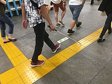 A visually-impaired person using a white cane is walking with assistance of the tactile pavement