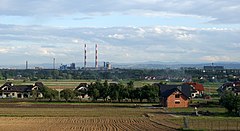 Fotografia barwna, wiejski krajobraz, na pierwszym planie zaorane pole i domy jednorodzinne, na dalszym planie z lewej zabudowania kombinatu metalurgicznego z dwoma okazałymi kominami jednakowej wysokości pomalowanymi w poziome czerwone i biale pasy