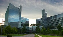 Two geometric all glass towers connected by a central atrium stand in front of a grassy walkway and under a dark and cloudy sky