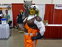 Femme maquillée en vert avec un costume marron, orange et blanc.