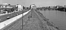 Schwarzweißfotografie in der Obersicht von einer Mauer mit Überkletterungsschutz, die sich von links unten zur Bildmitte erstreckt, und einem Fluss auf der rechten Seite. Zwischen Mauer und Fluss verläuft eine schmale Grünfläche mit Asphaltweg. Rechts am Ufer stehen Betonpfeiler. Links von der Mauer ist eine vielbefahrene Straße und im Hintergrund ist rechts von großen Gebäuden eine Brücke.