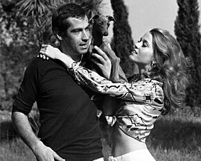 A man (Roger Vadim) standing with a dog on his shoulder with a woman (Jane Fonda) next to him.