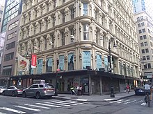 The lower floors of the Bennett Building, as seen from diagonally opposite the intersection of Fulton and Nassau Streets. The ground level contains darker-colored storefronts, while the other stories contain a light-colored facade.