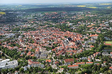 Barevná fotografie s leteckým pohledem na město s oválným historickým centrem uprostřed zalesněné krajiny