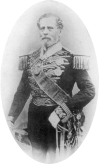 Tintype photograph depicting a full-length portrait of an older, bearded man wearing an elaborately embroidered military dress uniform with heavy epaulets, sash of office, sword, gloves and holding a bicorn hat in his right hand