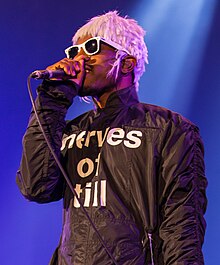 Artist performing onstage in a black jumpsuit reading "nerves of till", pink wig, white plastic sunglasses, and Converse-type sneakers.