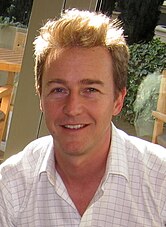 A Caucasian man with black hair and glasses. He is wearing a blazer and a shirt with the top button loosen.