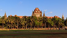 A brown building with a central tower and sloping roofs surrounded by trees. A grassy ground and a coconut tree are in front of it.