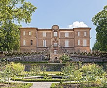 La photo couleur présente des terrasses fleuries au premier–plan et un château style classique en briques rouges.