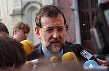 Photographie d'un homme brun à la barbe poivre et sel. Il parle devant de nombreux micros.