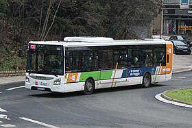 Photographie en couleurs d’un autobus blanc de type Iveco Urbanway avec des traits obliques bleu, orange et vert devant des arbres verts.