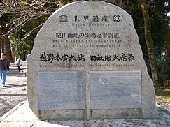 An image of people walking on a gravel forested path, the central focus is a large rock monument with a brass plate commemorating the establishment of the UNESCO World Heritage site.