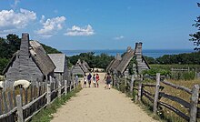 A modern-day photograph of a village consisting of small, primitive wooden houses. Most of the houses have thatched roofs. In the distance is a large expanse of ocean and a clear blue sky.