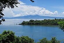 Photographie en couleur d'un rivage couvert de forêts avec derrière la mer. En arrière plan une grande montagne domine le paysage.