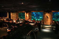 A photograph of the interior of a dimly lit restaurant where people are eating next to three windows into an aquarium housing a variety of marine organisms