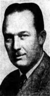 Black-and-white photographic portrait of a smiling man with slicked-back hair, a toothbrush mustache, and well-defined facial features. He eyes the camera with a classically handsome confidence and wears a suit.