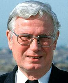 Colour news photo of a half-smiling, elderly, clean-shaven, white-haired man wearing spectacles.