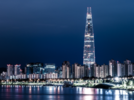 A nighttime shot of the Lotte tower from across the river. The tower is lit up and is far taller than any of the apartment buildings around it. By the riverside, a row of streetlights are shining on the river.