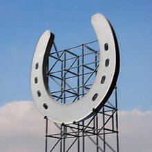 A digital image of a giant horseshoe suspended in front of metal scaffolding. A blue sky with low clouds appears in the background.