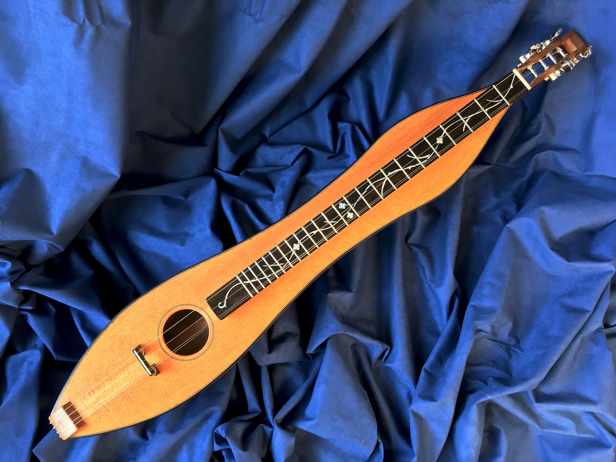 An hourglass-shaped mountain dulcimer with four strings, made from walnut, cedar and ebony, with mother-of-pearl fretboard inlays, against a folded blue fabric background