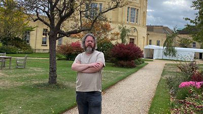 Johny Cassidy stands with his arms crossed on a gravel path looking into the camera. He’s got long grey hair and a full grey beard and is wearing a beige T shirt with black jeans and boots.