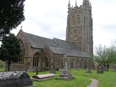 Gray stone building with ornate square tower and slate roof. In the foreground are gravestones.