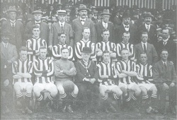 A football team comprising ten players in striped shirts and one in a shirt of a single colour pose for the camera. Five of the men are standing and the other six seated in front of them. Also posing with them are an elderly man in a bowler hat with a chain of office around his neck, and twelve men in business suits, some of whom are wearing hats. A crowd of spectators is visible behind the group.