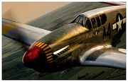 A close-up view of the front half of a single nosed-propeller aircraft is in mid-flight above land. The red nose faces left and the cockpit is in view from the top. The aircraft has dark propellers that are barely perceptible because of their high speed, gray base paint, and black letters reading: "A4". The word: "TUSKEGEE" is visible on the nose of the aircraft and other lettering and insignia can also be seen at higher resolutions. The "4" is followed by a United States roundel in black with a central white star.