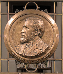 a circular bronze plaque has a raised impression of the bust of a man who is facing to the left. The place is located on bronze railings.