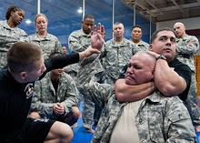 A man in fatigues rises his hand while another man in a black t-shirt holds his neck from behind with an arm to his neck and another to the back of his head. In the background men and women in fatigues observe.