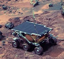 A Mars rover, topped with solar panels, on reddish-brown terrain.