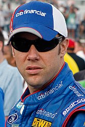 A man in his late thirties wearing a blue-and-white baseball cap and black sunglasses along with a blue jacket with sponsors' logos.