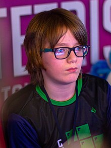 Headshot of Gibson facing slightly away from the camera to the right: wearing a green, blue, and black Tetris T-shirt and glasses