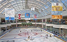 An ice hockey rink seen from a second storey above it. There are two teams playing at the far end. People are watching the game from both levels; there are stores behind them. Above the rink is a glass ceiling from which advertising banners hang promoting the tournament sponsors, as well as the Canadian and U.S. flags (June 27, 2015).