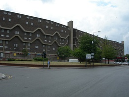 A long dark building with small windows and upper floors slightly overhanging lower ones. Two pale bands at the overhangs step up and down along the face of the building.